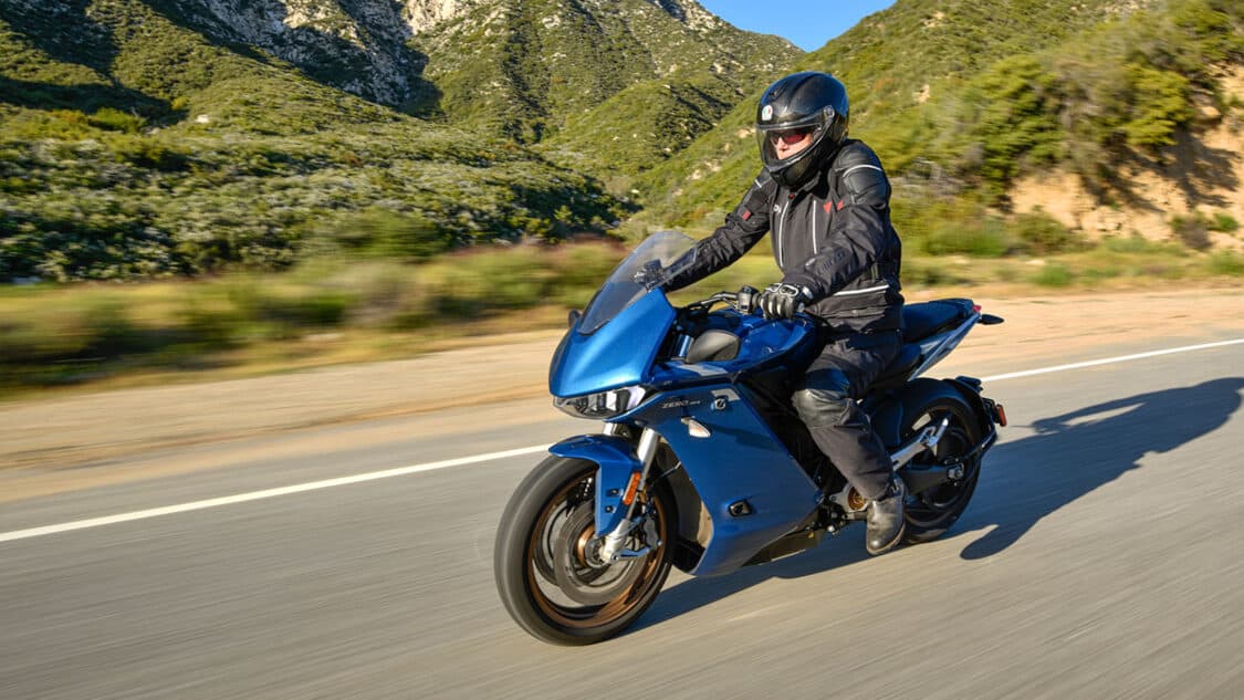 photo of man wearing helmet jacket and gloves riding an blue electric motorcycle on paved road near mountains