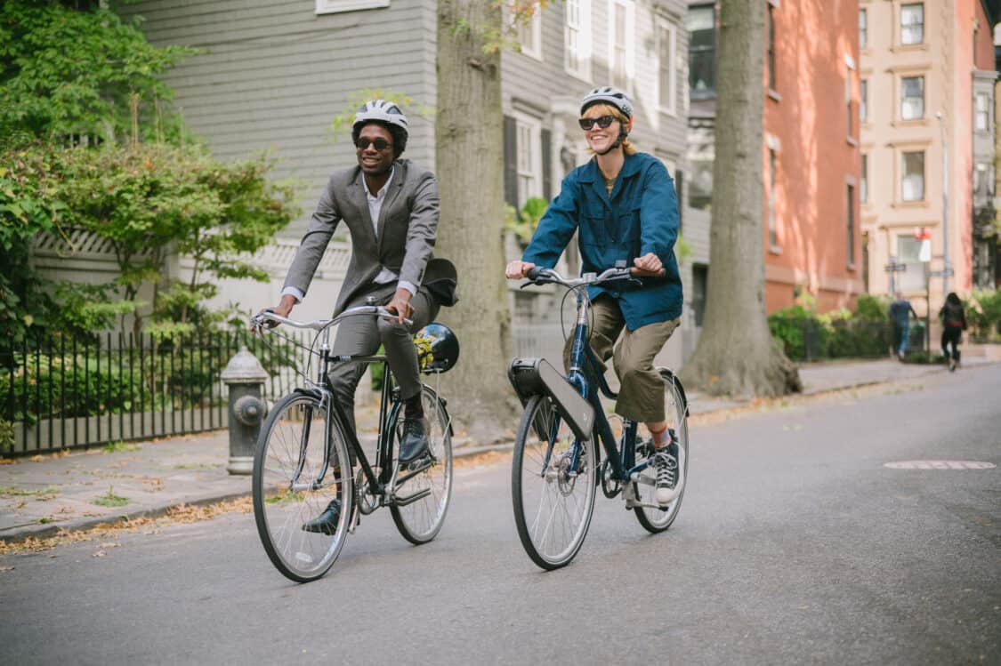 Photo of man and woman wearing helmets and riding bikes on a city street. The bike the woman is riding has the CLIP pedal-assist product installed. They are both smiling.