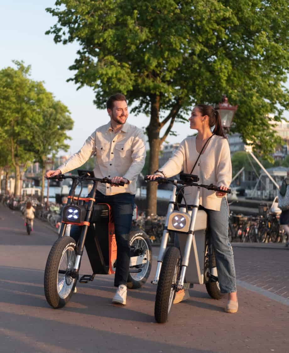 Photo of ASYNC A1 black and orange and silver electric bikes, man and woman standing on paved road in the city near the dock and boats.