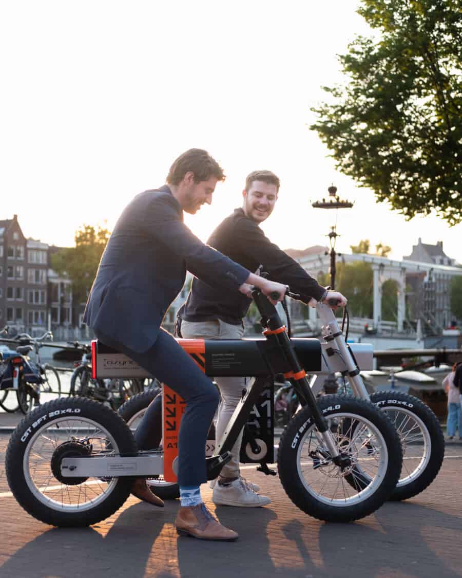 Photo of ASYNC A1 black and orange and silver electric bikes, men standing on paved road in the city near the dock and boats.