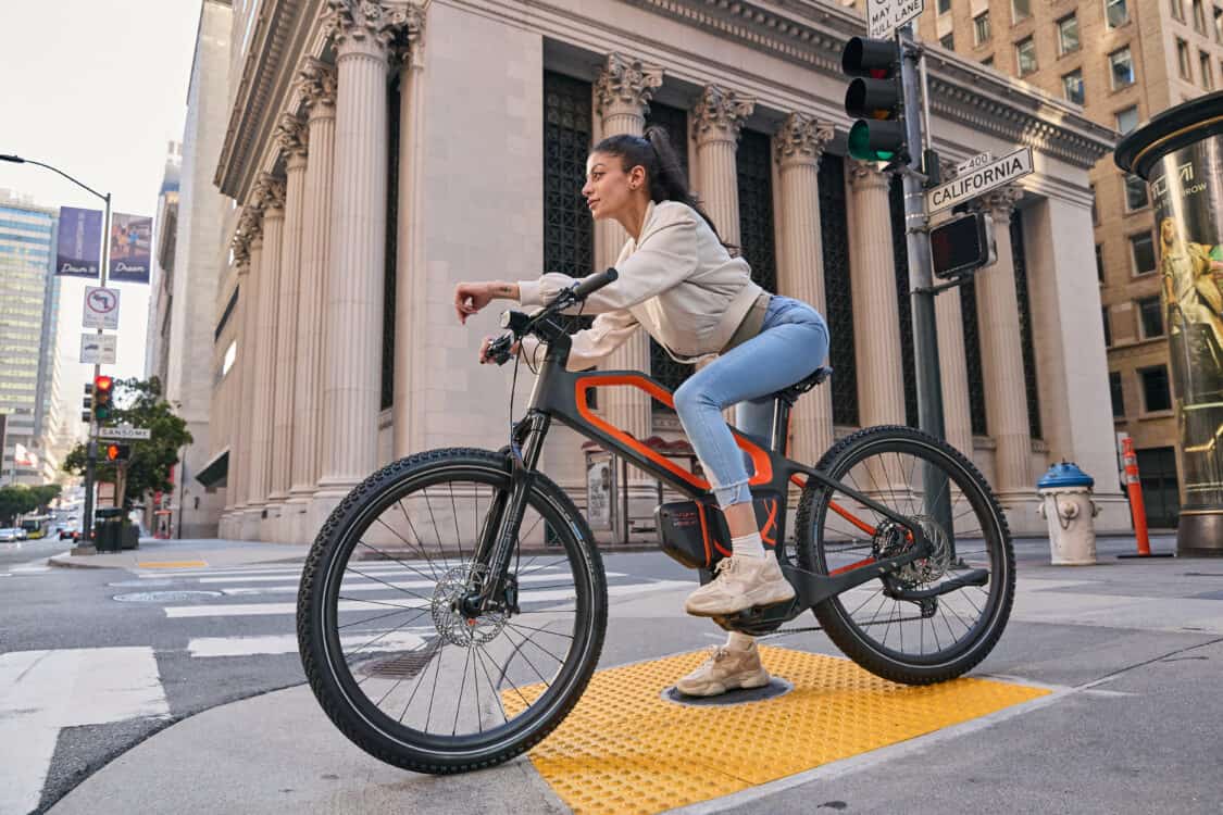 Photo of ASYNC H7 black and orange electric mountain bike, woman riding on sidewalk in the city