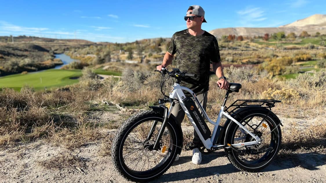 Senada fat tire electric mountain bike with man standing next to it with contrasting golf course and rough wild landscape surrounding