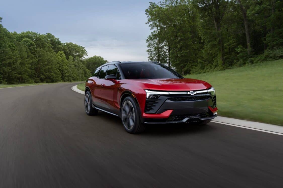 Passenger’s side view of the 2024 Blazer EV SS in Radiant Red Tintcoat driving on a road with trees.