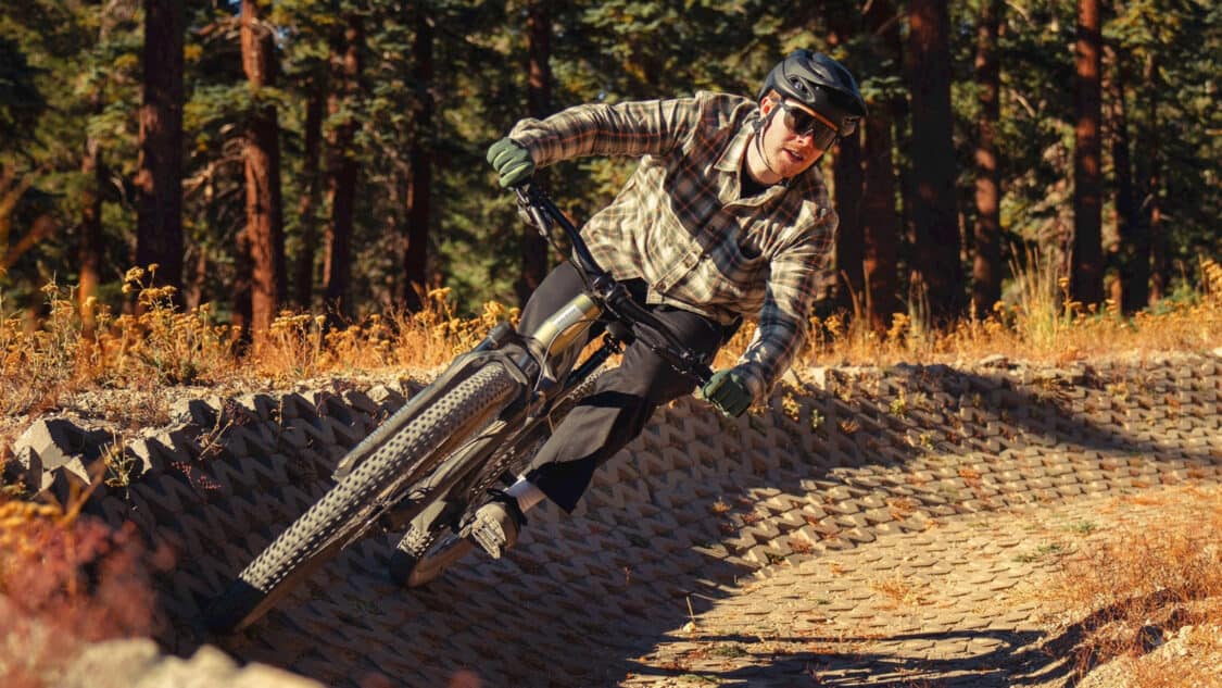 A mountain biker on the Aventon Ramblas electric mountain bike in a plaid shirt and helmet aggressively cornering on a cobblestone path, with a forest backdrop illuminated by sunlight