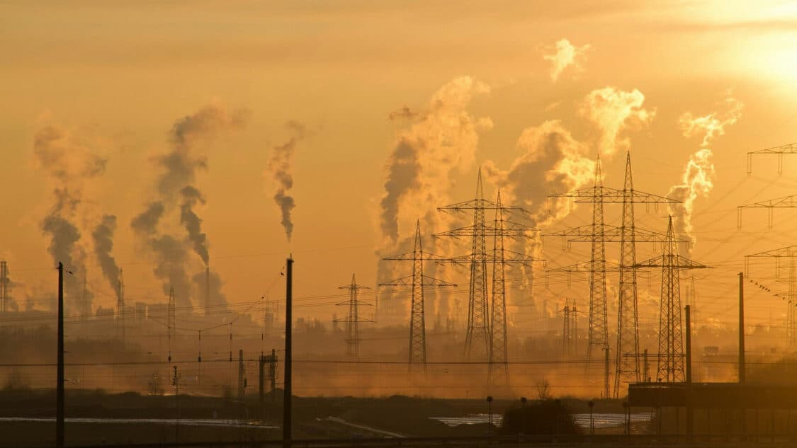 An industrial landscape at sunrise with multiple smokestacks emitting plumes of smoke into the air, power lines in the foreground, and a hazy sky, suggesting pollution and environmental impact.