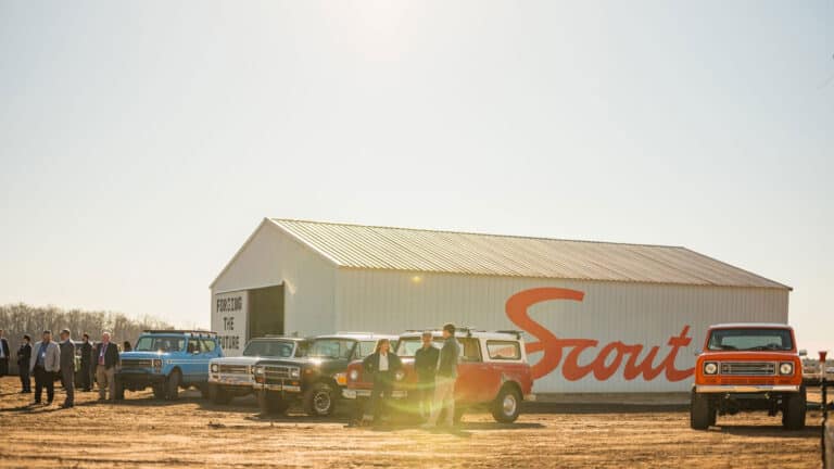 Scout Motors manufacturing plant groundbreaking ceremony with large shed with "Scout" written on the side and vintage vehicles parked around it.