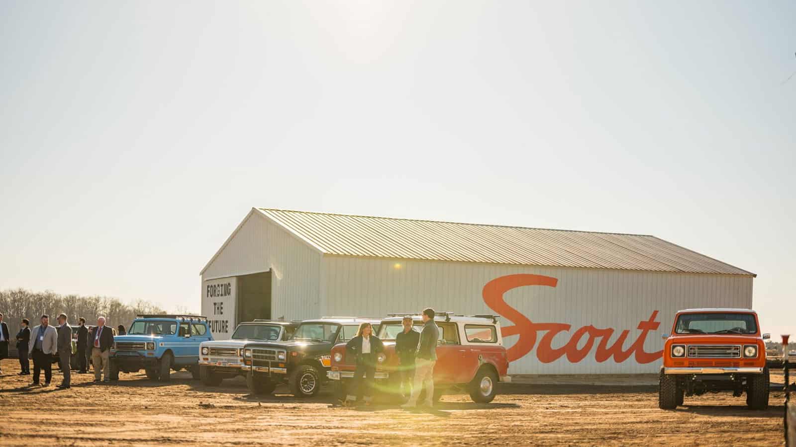 Scout Motors manufacturing plant groundbreaking ceremony with large shed with "Scout" written on the side and vintage vehicles parked around it.