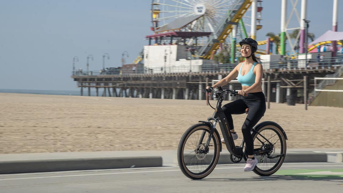 Woman riding AIMA Santa Monica commuter E-bike next to amusement park on beach