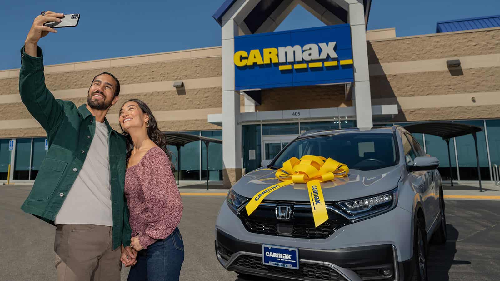 Carmax storefront with couple taking a selfie in front of an affordable car with a large yellow ribbon