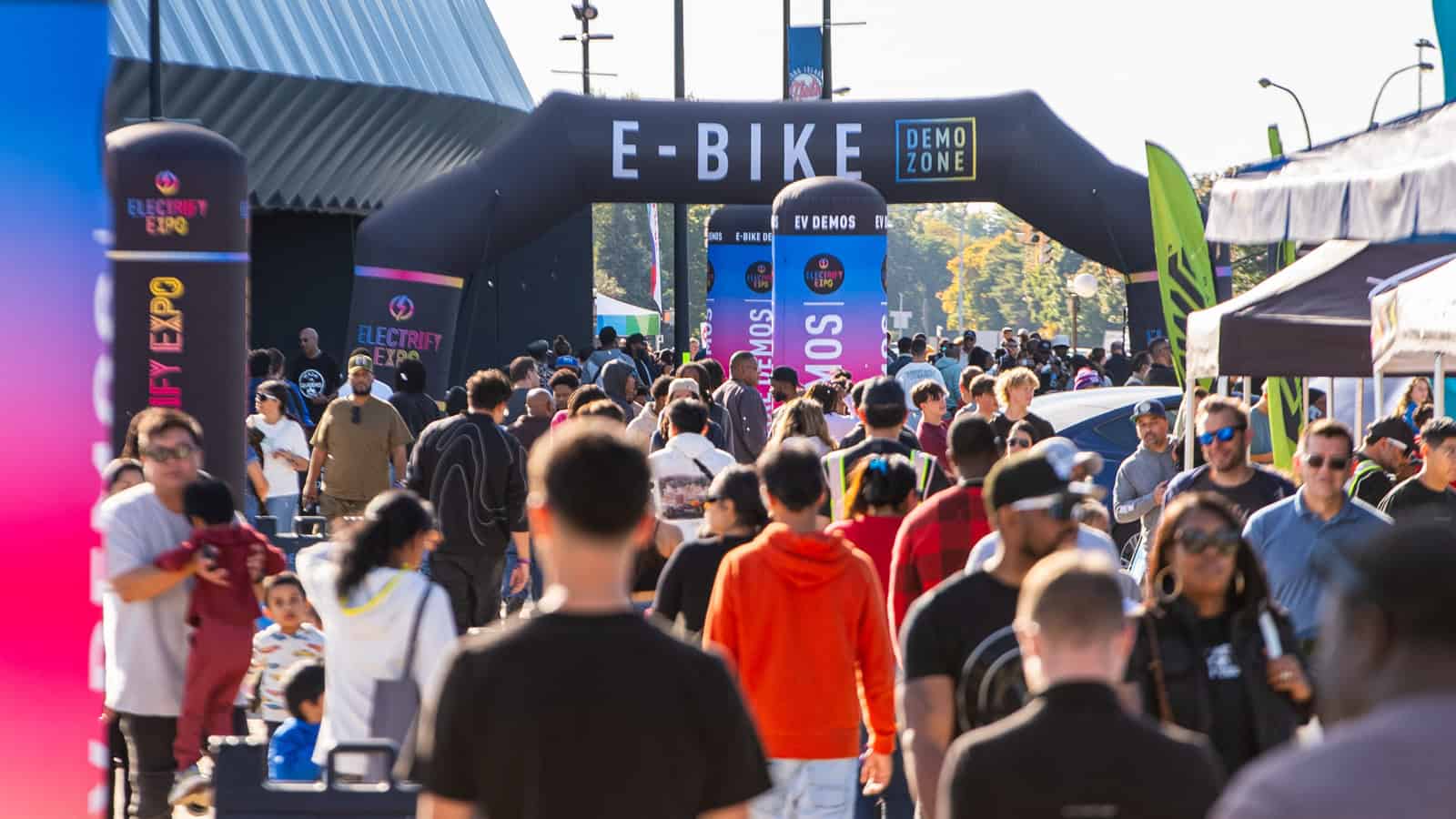 A bustling crowd at an outdoor Electrify Expo event featuring an E-Bike Demo Zone, showcasing local dealers, products, and demos for attendees of all ages.