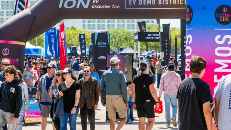 Crowds walk through the EV Demo District at Electrify Expo, surrounded by banners from major automakers.