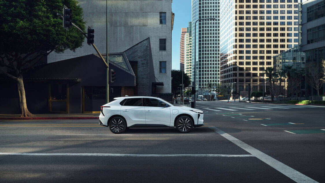 Side view of a white 2026 Toyota bZ electric SUV stopped at a city intersection, surrounded by tall office buildings.