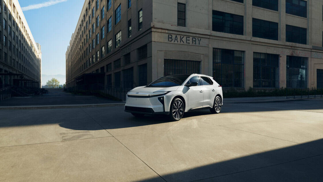 Front-side view of a white 2026 Toyota bZ electric SUV parked in an industrial alleyway between two large buildings marked “BAKERY.”
