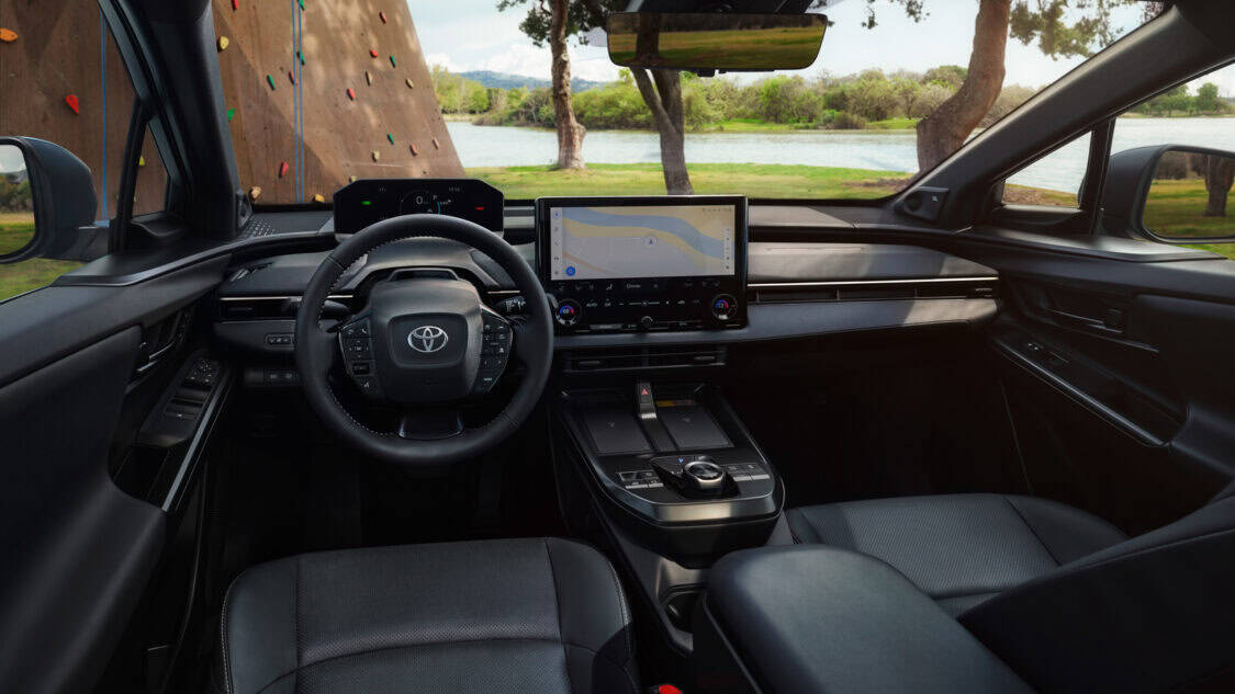Interior view of the 2026 Toyota bZ electric SUV showing the steering wheel, wide touchscreen display with navigation, and sleek dashboard design, with a lake and climbing wall visible outside.