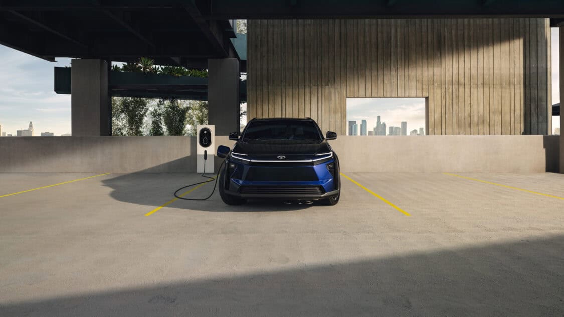 Front view of a blue and black 2026 Toyota bZ electric SUV charging at a station in a rooftop parking garage with a city skyline in the background.