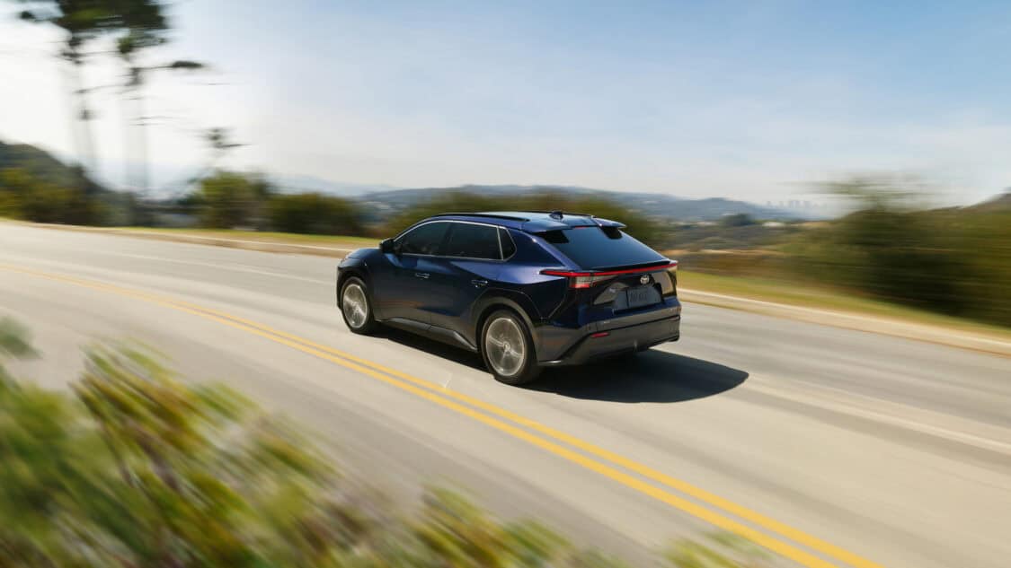 Rear view of a blue 2026 Toyota bZ electric SUV driving on a scenic road with hills and a distant city skyline in the background.