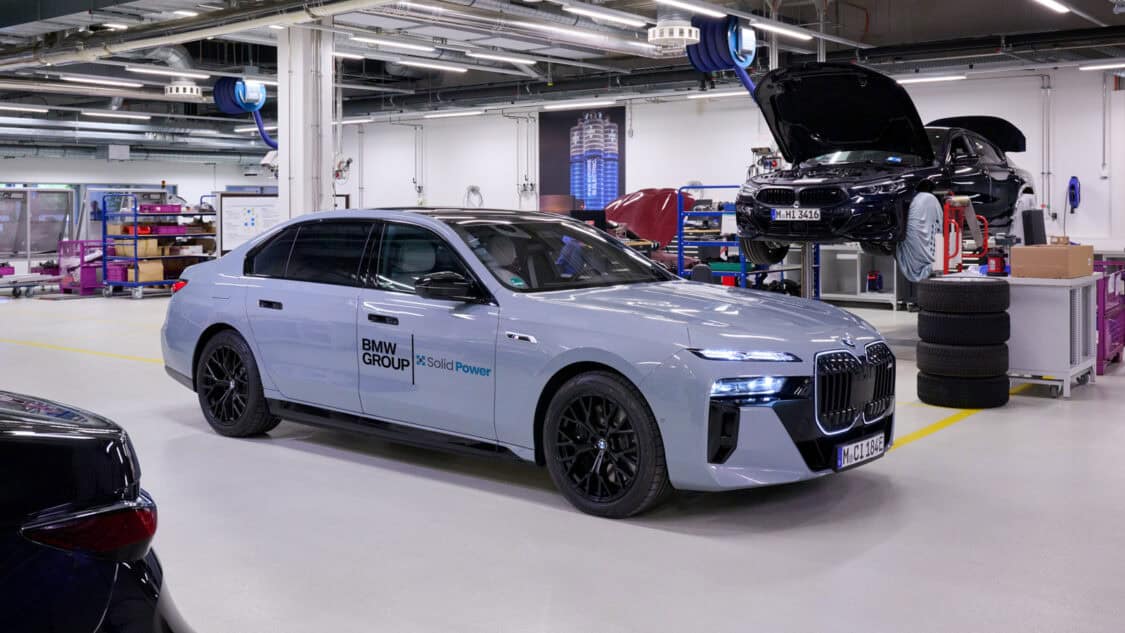 A BMW i7 test vehicle featuring Solid Power all-solid-state battery technology is parked inside a BMW Group workshop near other vehicles under maintenance.