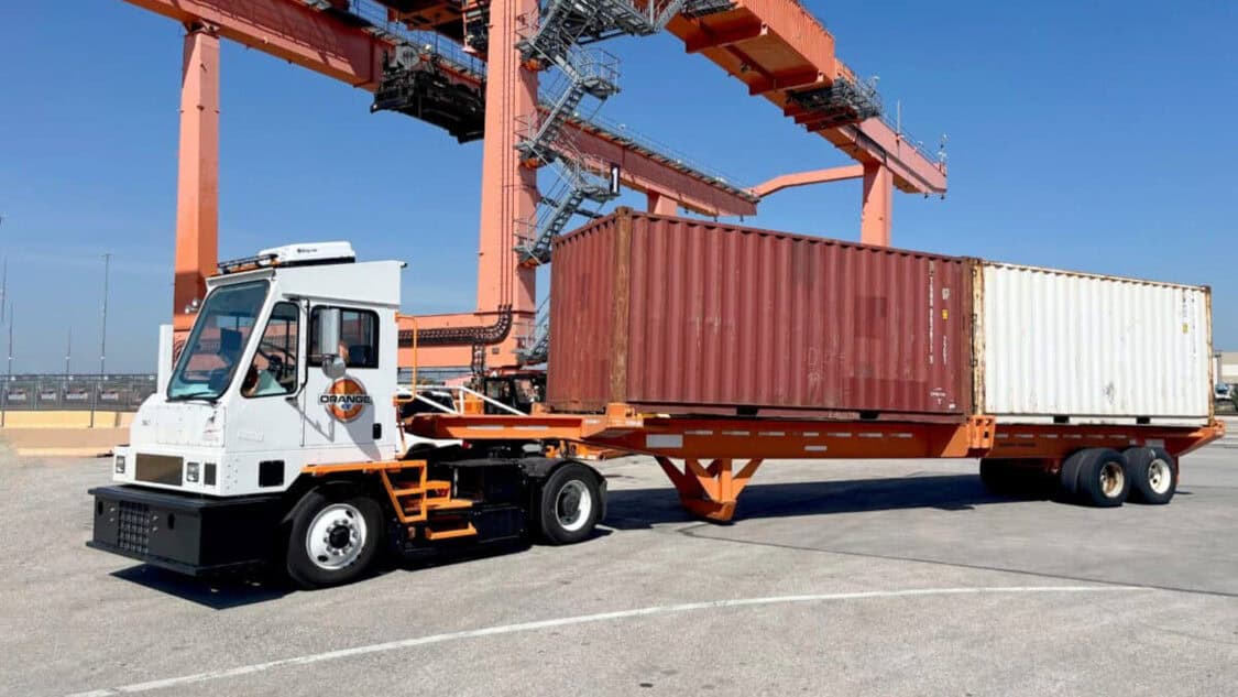 Orange EV electric yard truck hauling a chassis with shipping containers at an intermodal rail facility.