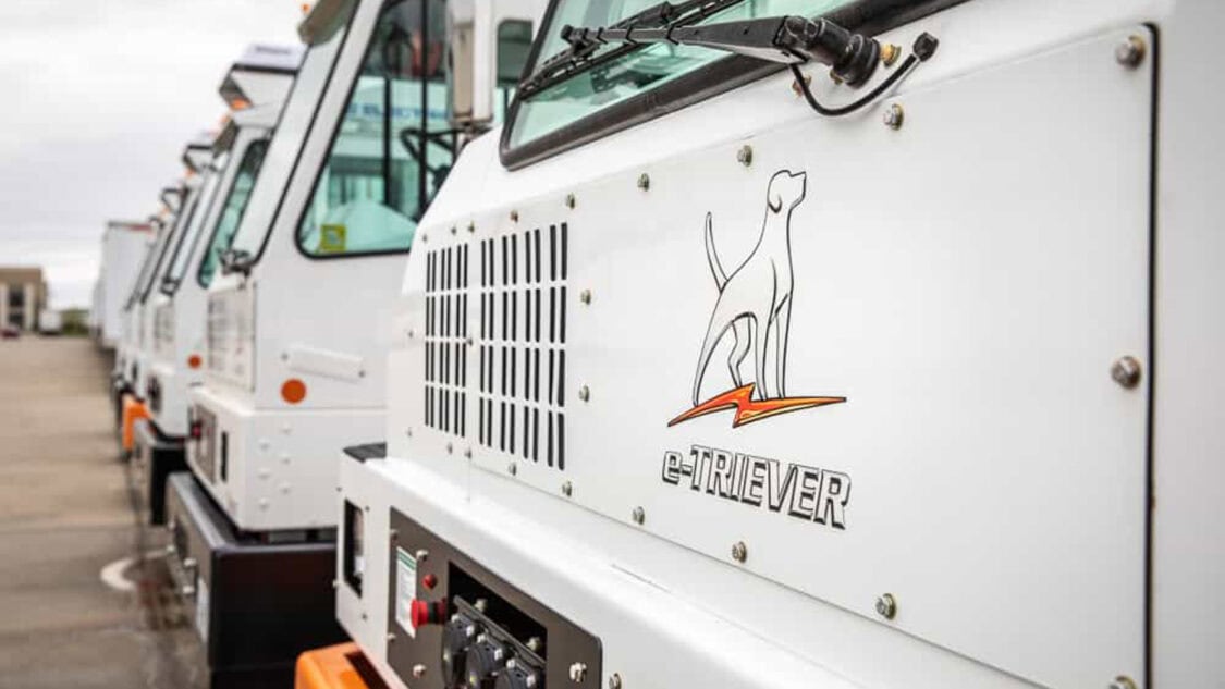 Close-up view of multiple Orange EV e-TRIEVER electric yard trucks lined up, highlighting the vehicle logo featuring a dog and lightning bolt.