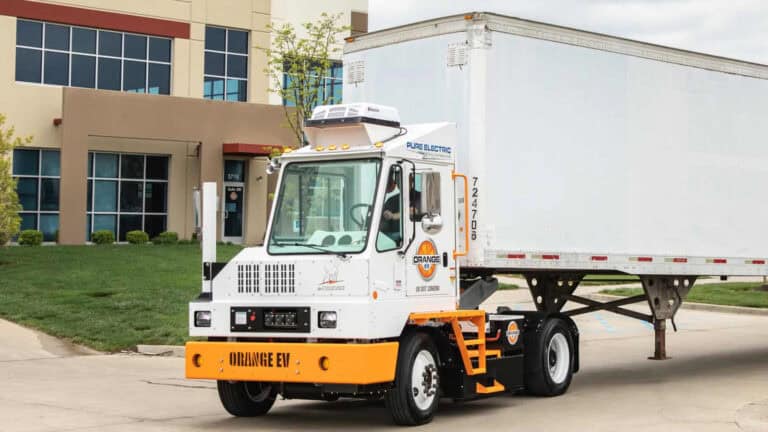 Orange EV pure electric yard truck moving a trailer in front of a commercial office building.