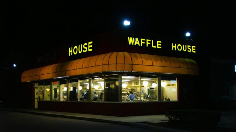 A Waffle House restaurant lit up at night, viewed from outside with people dining inside.