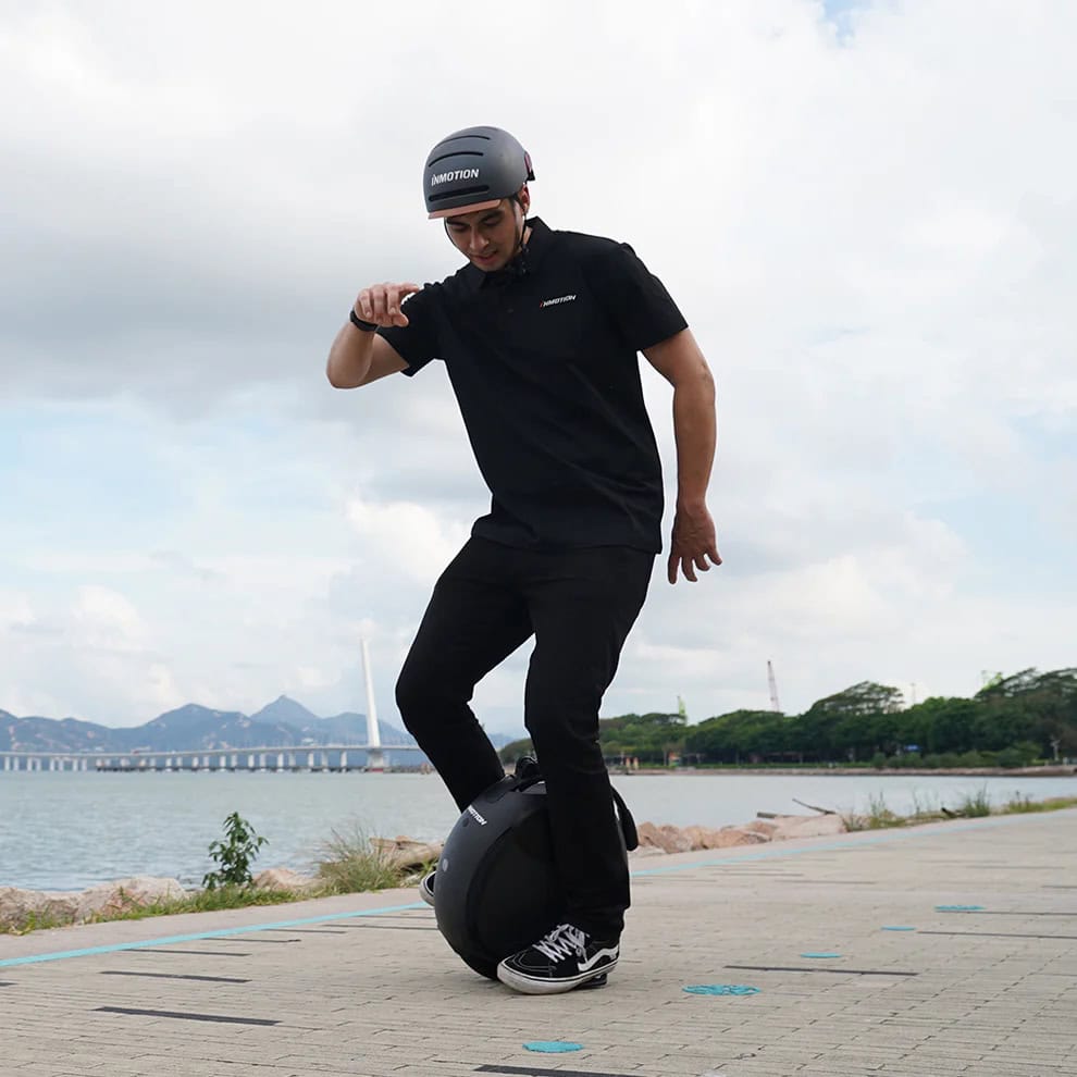 Rider balancing on an Inmotion V6 electric unicycle near a waterfront path, with mountains and a bridge visible in the background.