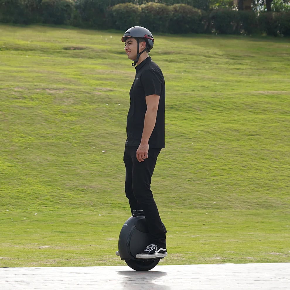 Person riding an Inmotion V6 electric unicycle on a paved path beside a grassy field, wearing a helmet and casual black outfit.
