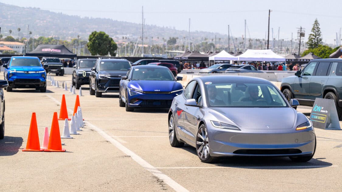 A line of electric vehicles, including a Tesla Model 3, Kia EV6, and other EVs, drives through EV Demo District at Electrify Expo event.