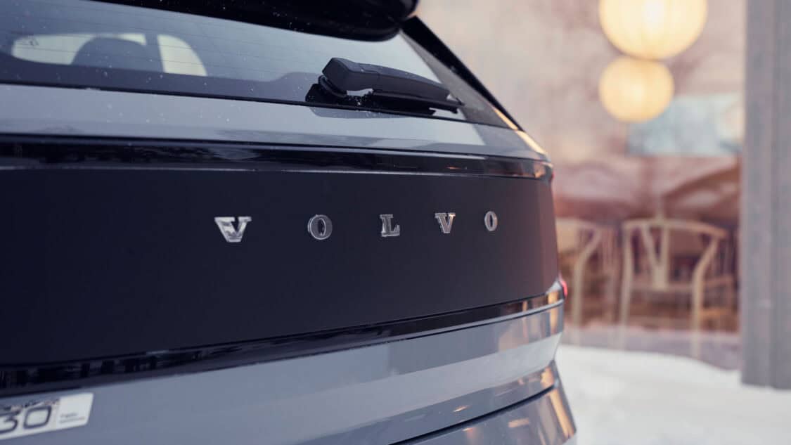 Close-up of the rear tailgate on the 2026 Volvo EX30 Cross Country featuring the Volvo lettering in chrome on a matte black panel with snowy surroundings and reflections of indoor lighting in the background.