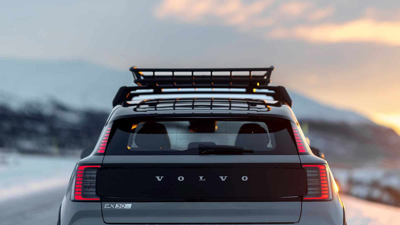 Rear view of the 2026 Volvo EX30 Cross Country with illuminated LED taillights and roof rack, parked on a snowy road at sunset with mountain scenery in the background.