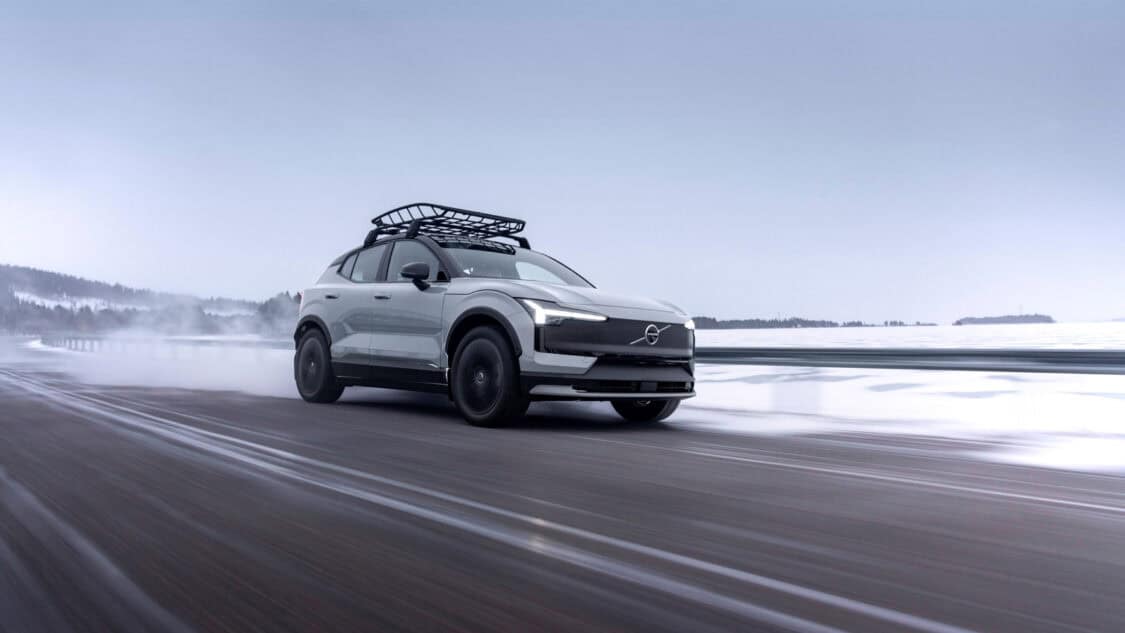 2026 Volvo EX30 Cross Country in motion on a wet, snowy road, featuring roof rack and blacked-out wheels, captured against a wintry landscape with overcast skies.