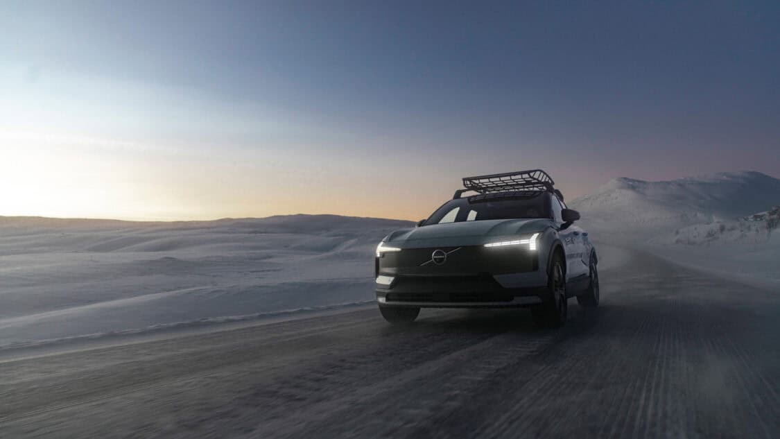 2026 Volvo EX30 Cross Country driving on a snow-covered road at dusk, with headlights on and roof rack installed, framed by snowy hills and a fading sunset sky.
