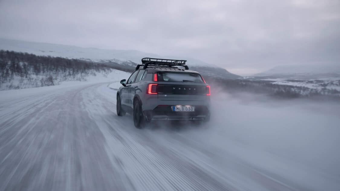 Rear view of the 2026 Volvo EX30 Cross Country driving on a snow-covered road with taillights illuminated, creating a snow trail behind, surrounded by snowy mountains and overcast skies.