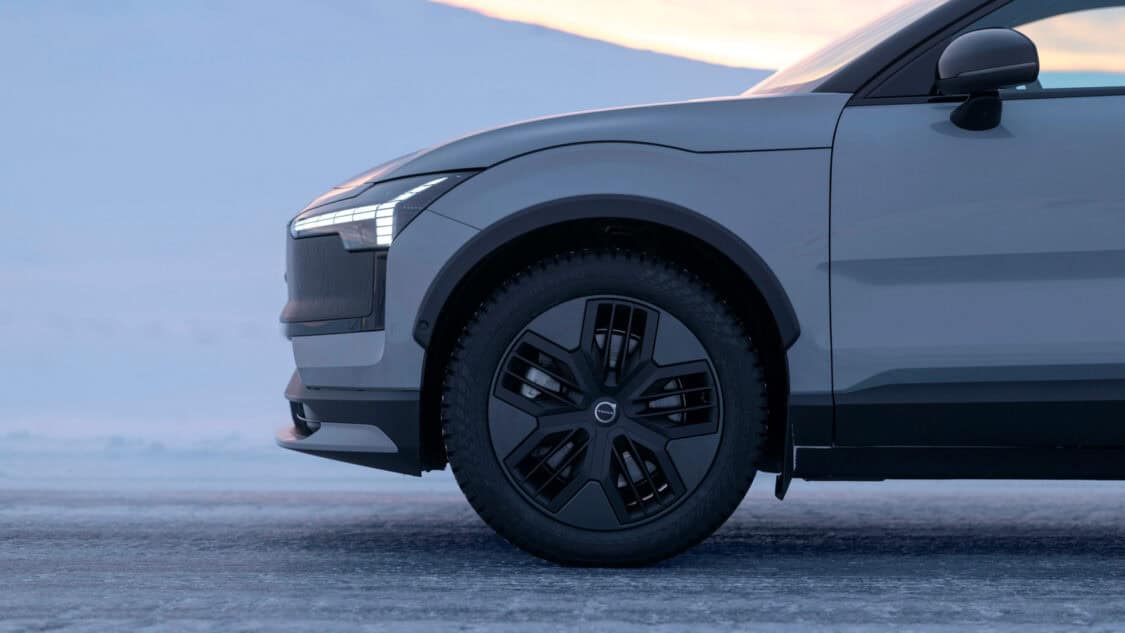 Side view of the front end of the 2026 Volvo EX30 Cross Country, highlighting the illuminated LED headlights, black fender trim, matte black alloy wheel, and winter tire on a snowy surface at sunset.
