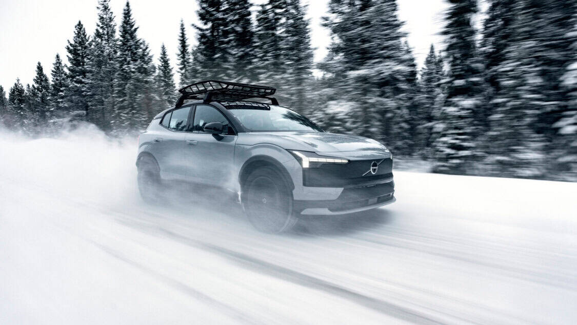 2026 Volvo EX30 Cross Country driving at speed through a snow-covered forest road, with roof rack installed and headlights on, kicking up snow behind the wheels.