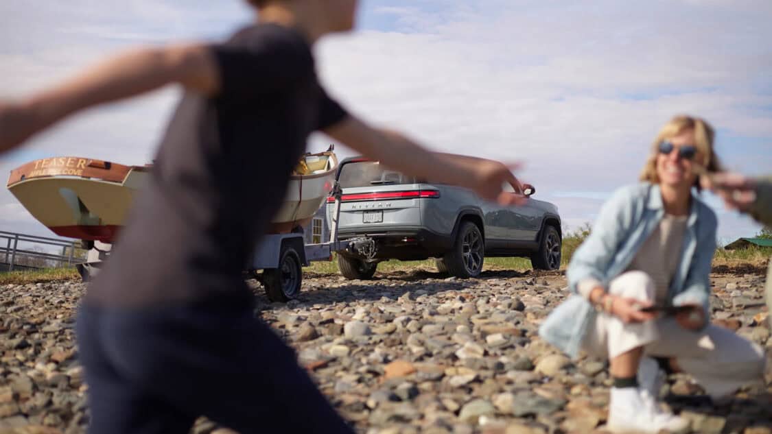 A Rivian truck parked near the beach in the Hamptons, towing a small boat, as a child runs past and adults relax on the rocky shore.