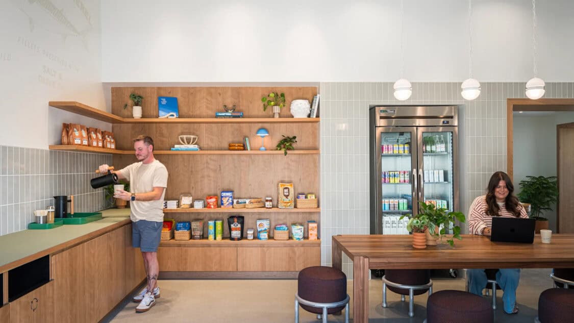 A man pours coffee beside shelves of curated snacks while a woman works on a laptop at a shared table inside Rivian’s Southampton Charging Outpost EV lounge.