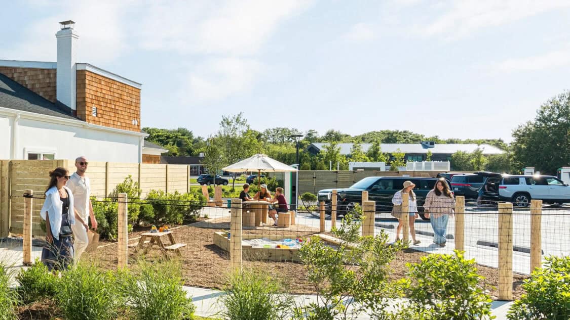 Families walk past Rivian’s Southampton charging outpost as others relax under umbrellas near a fenced play area and EVs charge in the background.
