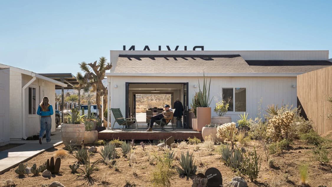 A visitor relaxes outside Rivian’s Joshua Tree charging outpost, surrounded by desert plants, with EV chargers tucked behind solar-covered parking.
