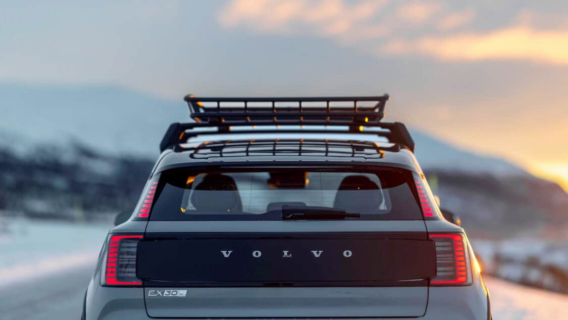 Rear view of the 2026 Volvo EX30 Cross Country with a roof rack, parked on a snowy road at sunset with mountains in the background.