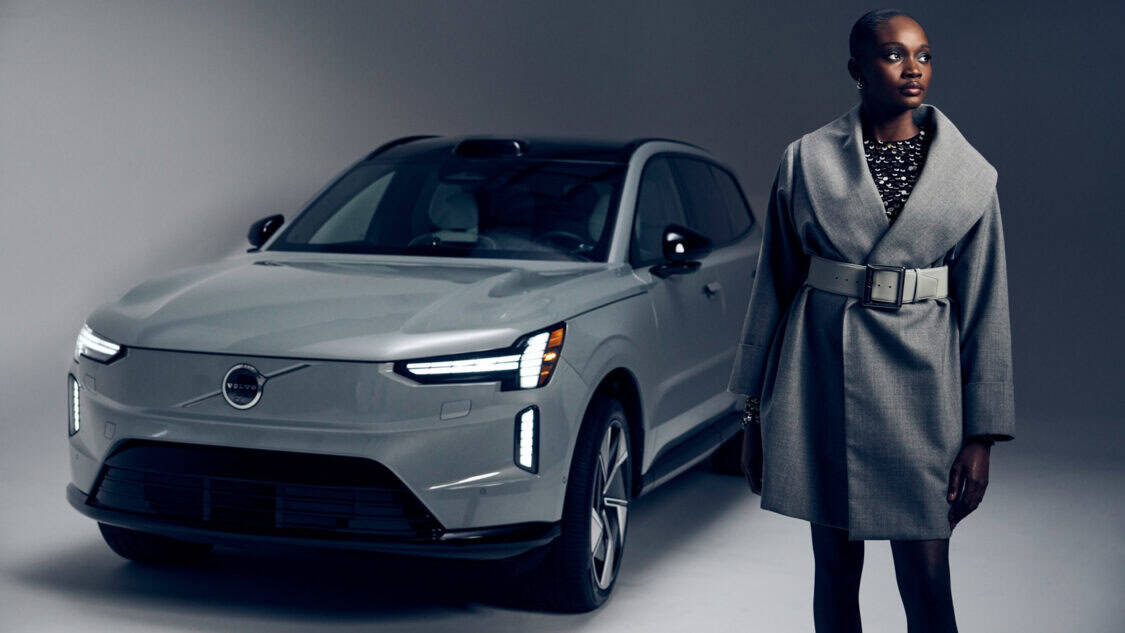A model wearing a grey Sergio Hudson car coat and Nordico belt stands beside the Volvo EX90, blending fashion design with electric luxury.