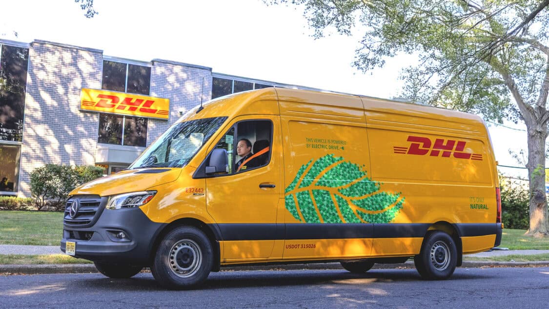 A yellow DHL Mercedes-Benz eSprinter electric van parked outside a DHL facility, highlighting its zero-emission delivery message and green leaf graphic.