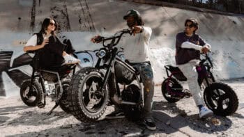 Three friends sit on their SUPER73-MZFT electric bikes near a graffiti wall, laughing and relaxing before a casual urban ride.