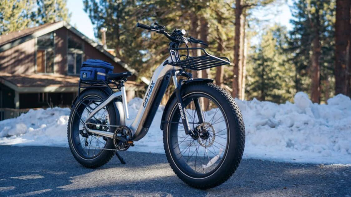A GOTRAX Rambo fat-tire e-bike parked on a snowy road near a cabin, highlighting its winter-ready frame, wide tires, and commuter rack for holiday riders