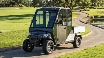 Massimo MVR HVAC electric utility vehicle with enclosed cab and cargo bed driving on a landscaped campus road for fleet and commercial use.
